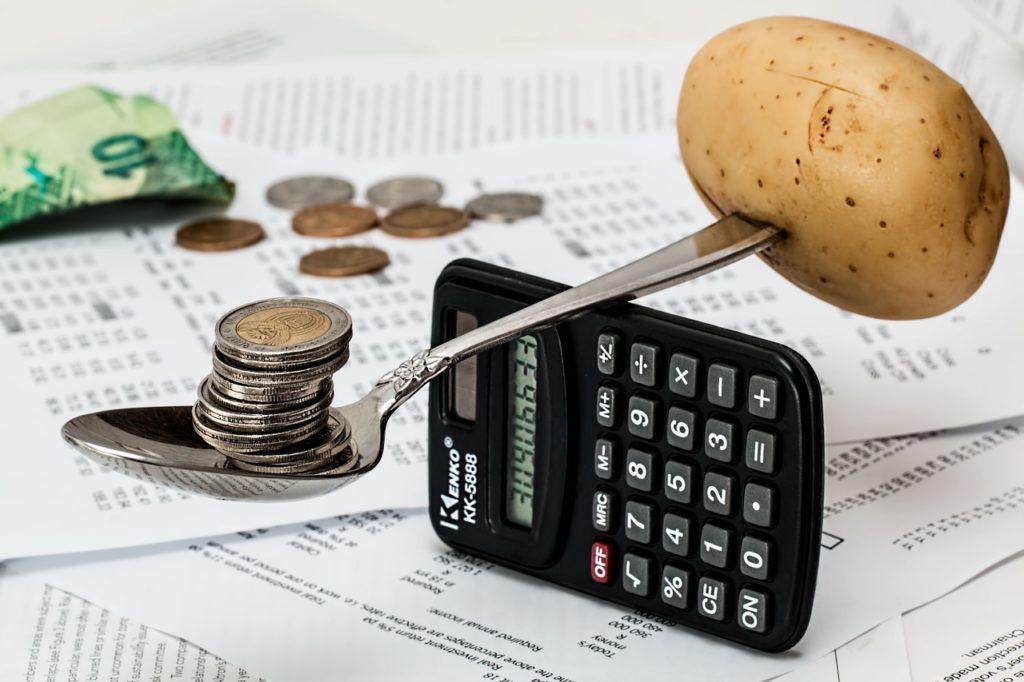 Calculadora com uma colher em cima servindo de balança entre moedas e uma batata. A imagem ilustra post sobre orçamento escolar