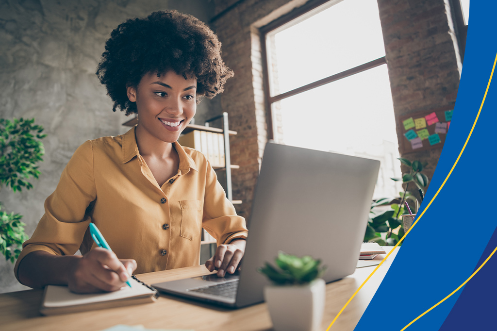 Mulher sentada em frente a um notebook e, com uma das mãos, fazendo anotações em um caderno. A imagem ilustra post sobre a organização de tarefas do fim do período letivo.